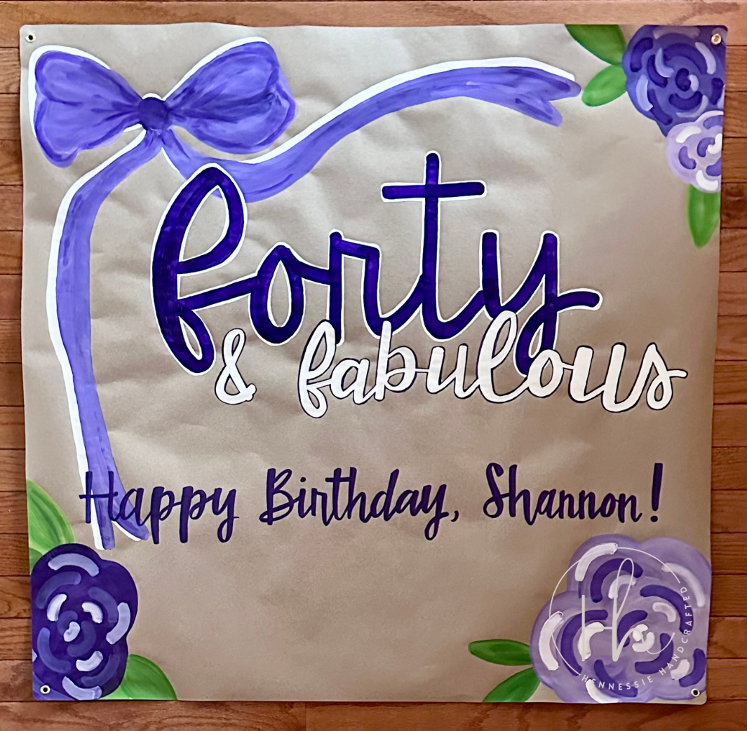 A custom hand-painted banner for a 40th birthday featuring purple roses, a purple ribbon graphic, and the text 'Forty & fabulous Happy Birthday, Shannon!' on a beige background, displayed on a wooden surface.