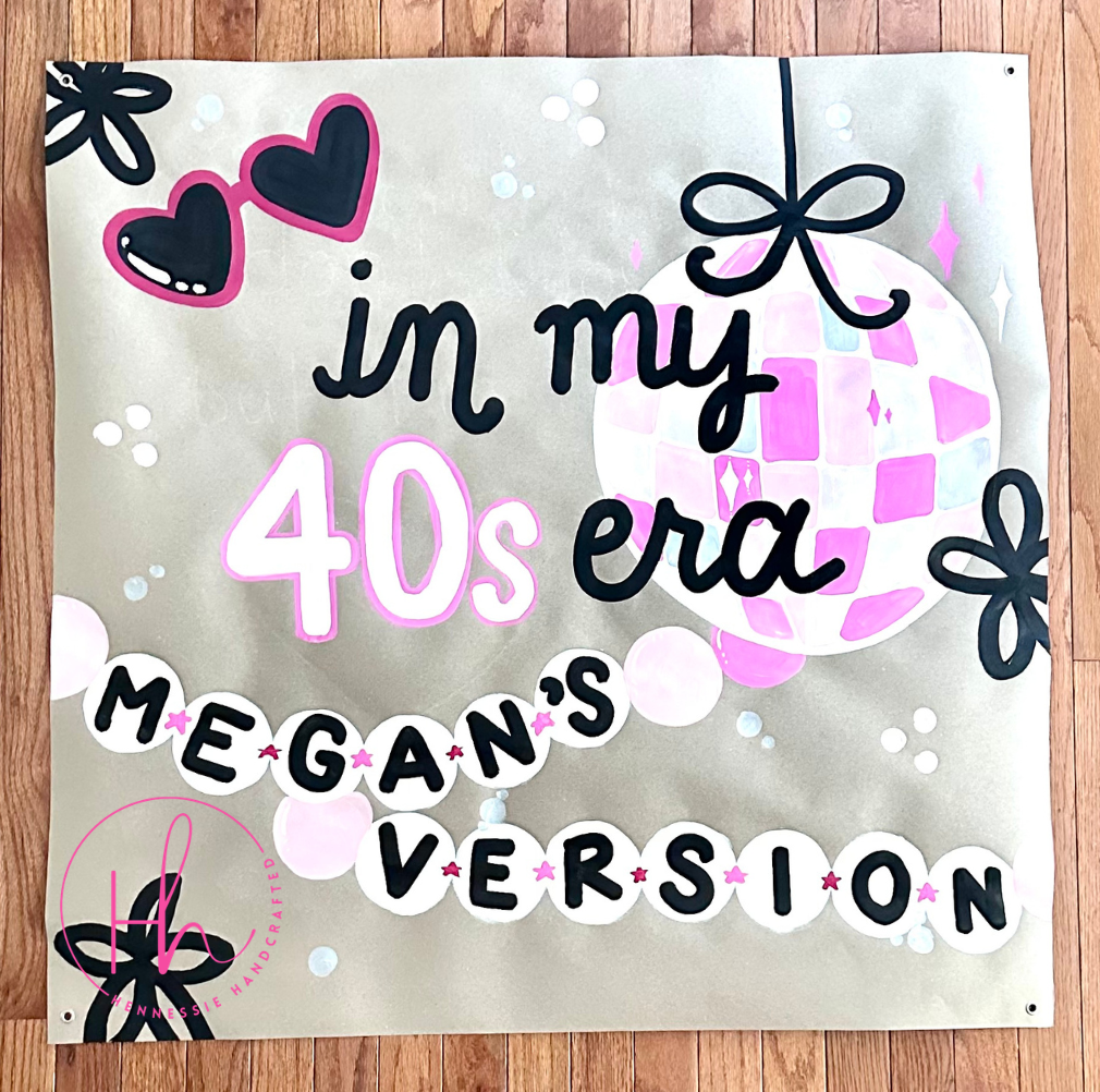A square printed banner or sign laid flat on a wood floor, featuring the text 'in my 40s era MEGAN'S VERSION' with pink and black graphics including sunglasses, a disco ball, and floral motifs.