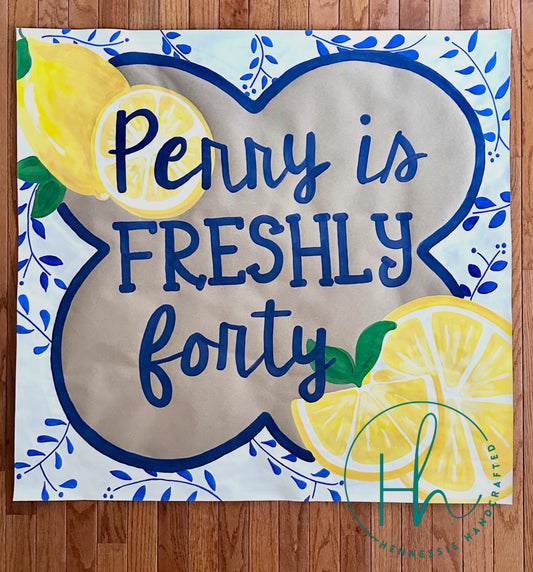 A square decorative sign or print with the phrase 'Penny is FRESHLY forty' in dark blue script and block lettering, surrounded by illustrated lemons and blue floral vines on a white background, photographed flat on a wood floor.