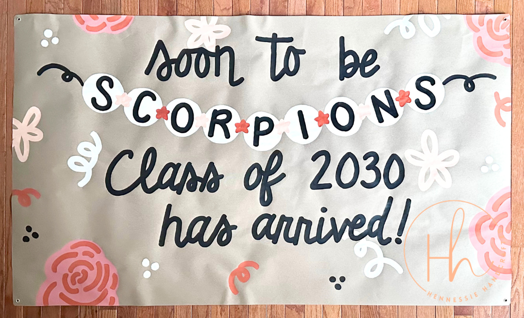 A rectangular banner with the text 'soon to be SCORPIONS Class of 2030 has arrived!' in black script, decorated with pink and white floral illustrations on a light beige background, displayed flat on a wooden floor.