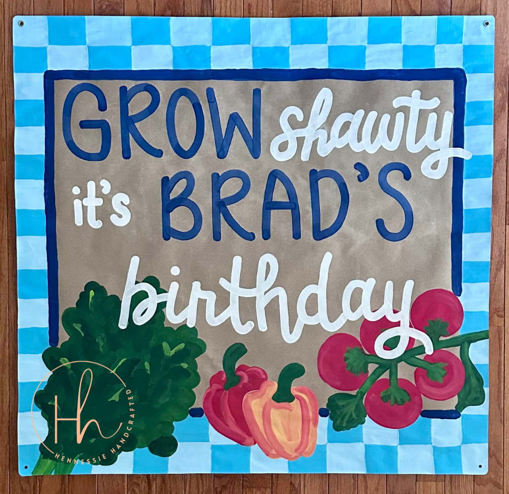 A square personalized garden sign with a blue and white checkered border, featuring the text 'GROW shawty it's BRAD'S birthday' in blue and white lettering on a tan background. The bottom is decorated with painted illustrations of green kale, a red bell pepper, and red tomatoes.