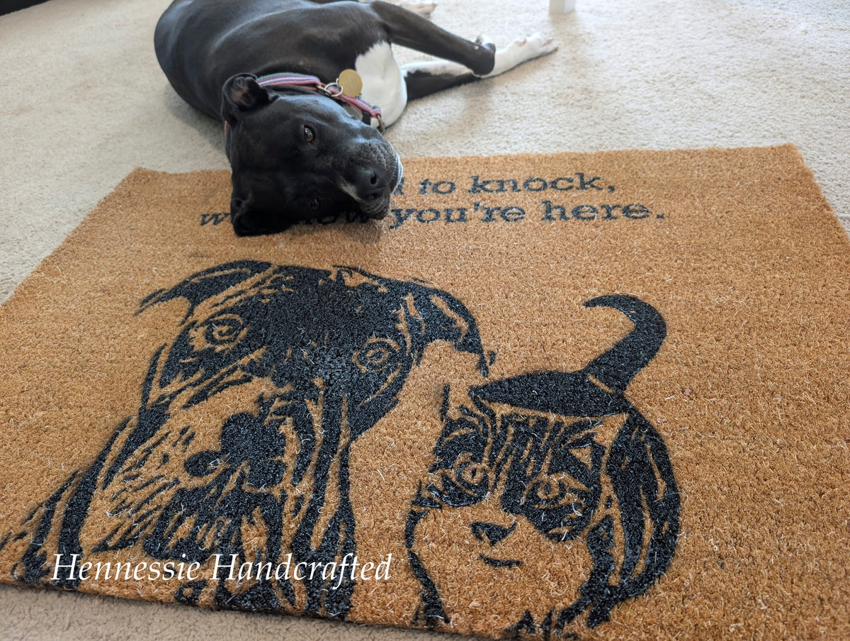A black dog lies on a tan coir doormat with a black printed illustration of two dogs and the text 'I love to knock, when you're here.' The photo is taken indoors on a carpeted floor.