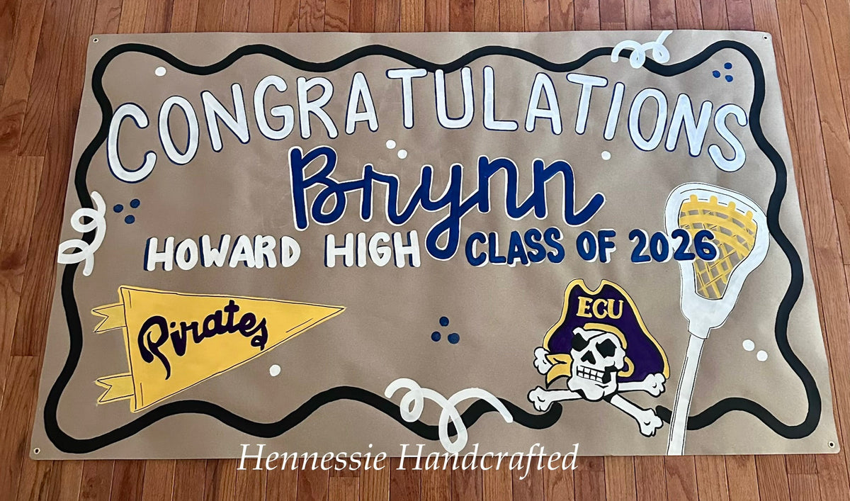 A handcrafted rectangular banner laid flat on a wooden floor, featuring the text 'CONGRATULATIONS Brynn HOWARD HIGH CLASS OF 2026' with decorative sports-themed graphics including a yellow pirate flag, a lacrosse stick, and an ECU skull-and-crossbones logo.
